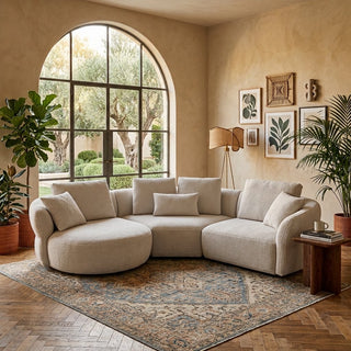 A curved modular sectional sofa in a warm cream or light beige fabric, styled in a beautifully decorated bohemian living room with an arched window, patterned rug, wooden floor, and potted plants.