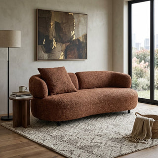 A curved, push back sofa upholstered in a textured rust-brown boucle fabric, styled in a modern living room setting with a matching throw pillow, a side table, a floor lamp, and a large abstract painting on the wall.