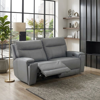 A grey reclining loveseat sofa shown in a modern living room setting with a window, floor lamp, shelving, and rug.