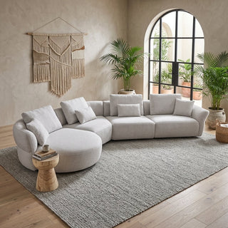 A light beige modular sectional sofa with a round chaise lounge section, styled in a bright, modern living room with a textured gray area rug, potted plants, and a macrame wall hanging.