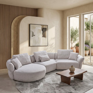 A light gray modular curved sectional sofa with matching throw pillows, styled in a modern beige living room with a wooden slat accent wall, abstract art, a wooden coffee table, and a gray patterned rug.