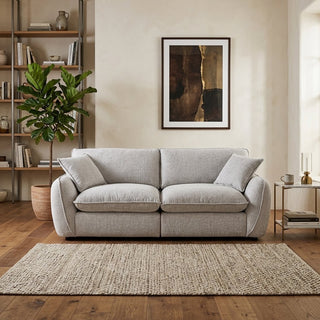 A light gray upholstered 2 seater sofa with two matching throw pillows, placed in a styled living room with a woven area rug, wooden floor, shelving, a potted plant, and framed wall art.