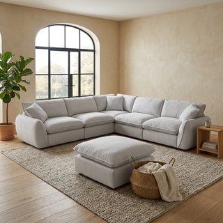 A light gray upholstered sectional sofa with matching ottoman in a styled living room with beige walls, wood floors, a large arched window, a potted plant, and a small side table.