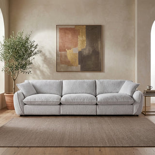 A light gray upholstered three-seat sofa in a styled living room setting, positioned on a textured brown rug beneath a framed abstract painting. A potted olive tree stands to the left, and a small side table with a book is on the right.