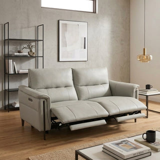 A light grey leather reclining loveseat sofa shown in a modern living room setting with its footrest extended, next to a black metal shelving unit and a side table.