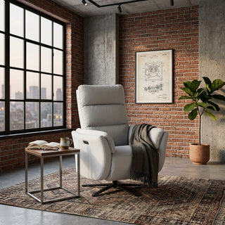 A light grey recliner sofa single with a dark gray throw blanket, set in a loft-style room with a brick wall, large window, and a potted plant.