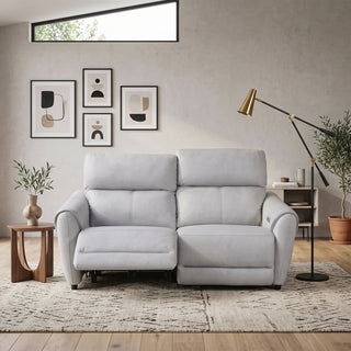 A light grey two-seater reclining sofa placed in a styled living room with framed wall art, a floor lamp, side table, plants, and a patterned rug.