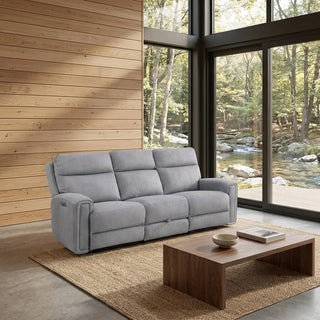 A light grey upholstered reclining loveseat sofa placed in a modern living room with wood-paneled walls, large floor-to-ceiling windows, a wooden coffee table, and a woven rug. A forest stream is visible outside the windows.