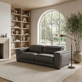 A modern dark grey leather recliner sofa placed in a styled living room with a large beige rug, built-in wooden shelves, a fireplace, an arched window, and a potted tree.