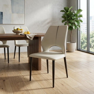 A modern dining room scene featuring a cream upholstered side chair with dark tapered legs in the foreground, alongside a wooden dining table and matching chairs. The room has light wood floors, a large potted plant, and a window looking out onto a city view.