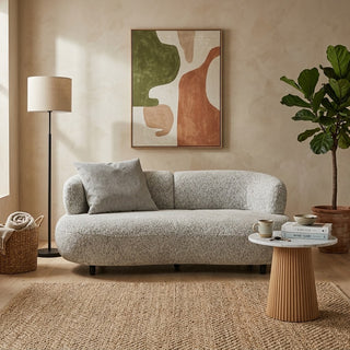 A modern push back sofa in a textured light gray boucle fabric, styled in a warm, boho-chic living room with a beige plaster wall, abstract art, a floor lamp, and a potted fiddle-leaf fig plant.