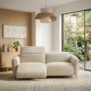 A modern two-seater electric recliner sofa in a warm off-white leather, styled in a minimalist living room with a light wood cabinet, a hanging pendant lamp, and large glass doors opening to a lush garden.