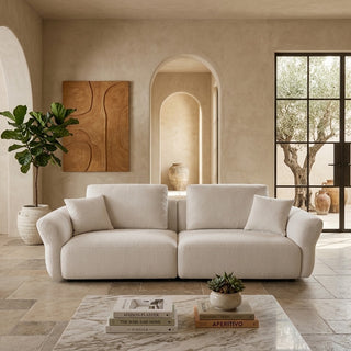 A modern two-seater sofa upholstered in a warm off-white textured fabric, styled in a minimalist, warm-toned Mediterranean living room with beige walls, a marble coffee table, and a large potted plant.