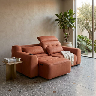 A rust-orange sofa sliders with a chaise lounge, styled in a modern living room with a textured beige wall, terrazzo floor, and a potted fiddle-leaf fig plant.