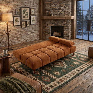 A tufted chaise folding sofa bed in a warm rust-brown velvet fabric, styled in a rustic living room with a green patterned rug, stone fireplace, and a snowy window view.