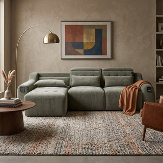 An olive green modular sofa sliders with a chaise lounge, styled in a modern living room setting with a patterned rug, a wooden coffee table, a gold floor lamp, and a framed abstract art print on the wall.
