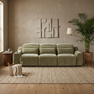 An olive green sofa sliders with matching cushions, styled in a modern living room setting with a woven rug, wooden floor, side table, lamp, and potted plant.