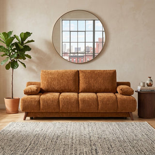 An orange-brown upholstered folding sofa bed in a styled living room setting with a potted plant, a round wall mirror, and a textured rug.