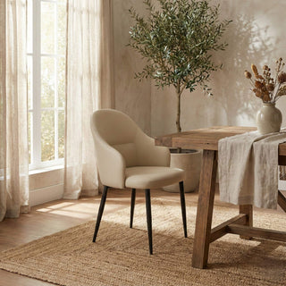 Beige chair in a room with a wooden table, plant, and curtains.