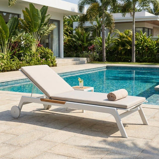 Beige lounge chair with wooden armrest on a patio by a pool, with a modern house and palm trees in the background.