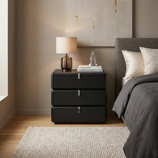 Black bedside table with lamp and books next to a bed in a bedroom.