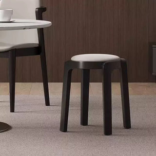 Black wooden stool with a white cushion on a gray floor, with a wooden wall and part of a table in the background.