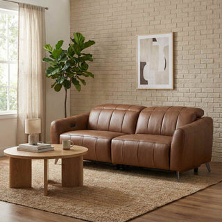 Brown leather sofa in a living room with a round wooden table, plant, and framed artwork on a brick wall.