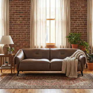 Brown leather sofa in a living room with brick walls and large windows.