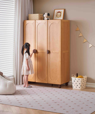 Child in a white dress standing next to a wooden childs wardrobe in a room with a light-colored rug and decorative items.