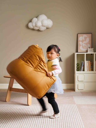 Child playing with a mustard-colored bean bag chair in a room with a bookshelf and decorative cloud on the wall.