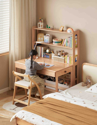 Child sitting at a wooden desk in a bedroom with books and toys.