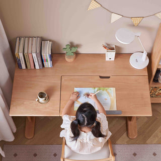 Child sitting at a wooden desk reading a book in a cozy room.