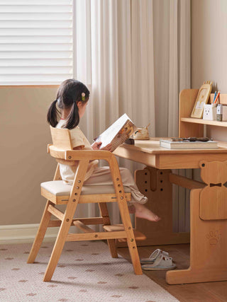 Child sitting at a wooden desk reading a book in a room with beige walls and curtains.