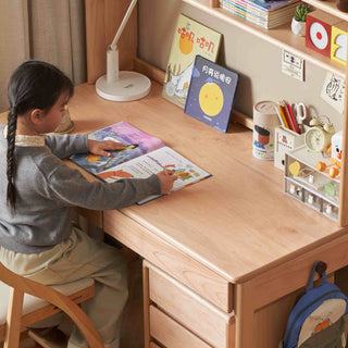 Child sitting at a wooden desk reading a book with various items on the desk.