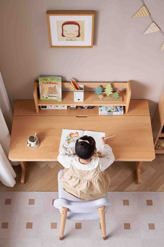 Child sitting at a wooden desk with a bookshelf, drawing on a piece of paper.
