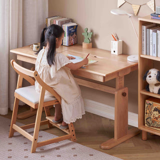 Child sitting at a wooden desk with books and a lamp, in a home study setting.