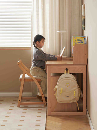 Child sitting at a wooden kids study table with a backpack, reading a book in a room with curtains.