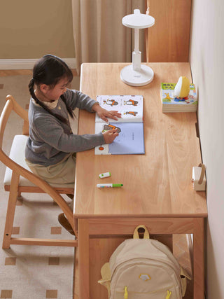 Child sitting at a wooden kids study table with a book and backpack, illuminated by a desk lamp.