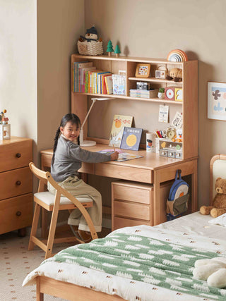 Child sitting at a wooden study desk in a room with a bed and shelves.