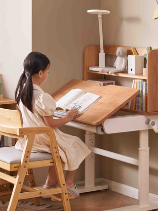 Child sitting at a wooden study desk with books and a lamp in a home setting.