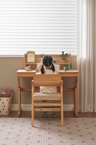 Child sitting at a wooden study table in a room with a window and curtain.
