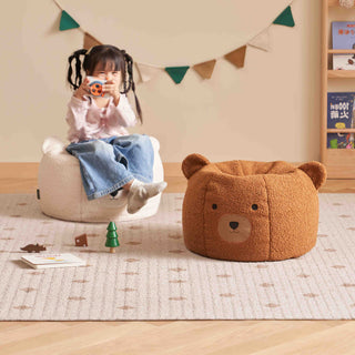 Child sitting on a bean bag chair next to a bear-shaped ottoman in a room with a wooden floor and colorful flags.