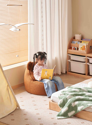 Child sitting on a bean bag chair reading a book in a cozy room with a bed and shelves.