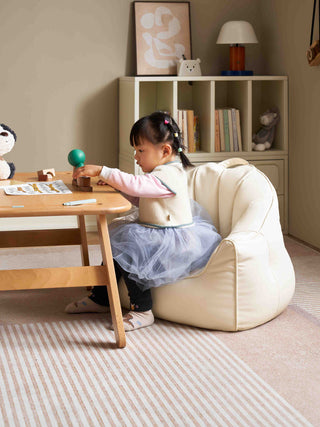 Child sitting on a bean bags for kids at a small wooden table in a room with a bookshelf and toys.
