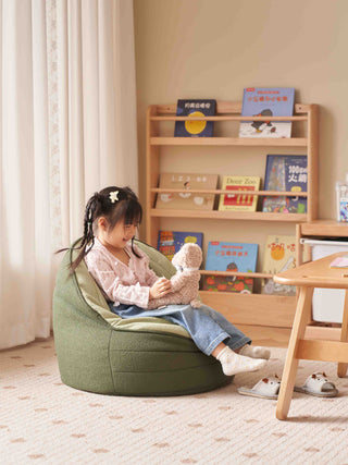 Child sitting on a green bean bag chair holding a teddy bear in a room with a bookshelf.