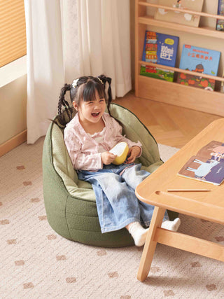 Child sitting on a green bean bag chair in a room with books and a table.