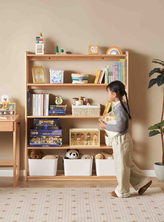 Child standing in front of a childrens wooden bookshelf filled with books and toys in a room.