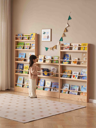 Child standing in front of a wooden bookshelf filled with books in a room.