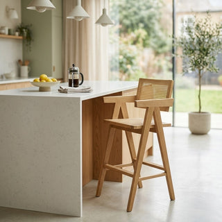 Modern kitchen with a white island and rattan bar stool, featuring a French press and fruit bowl.