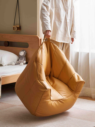 Person holding a mustard yellow bean bag chair in a room with a bed and stuffed animal.